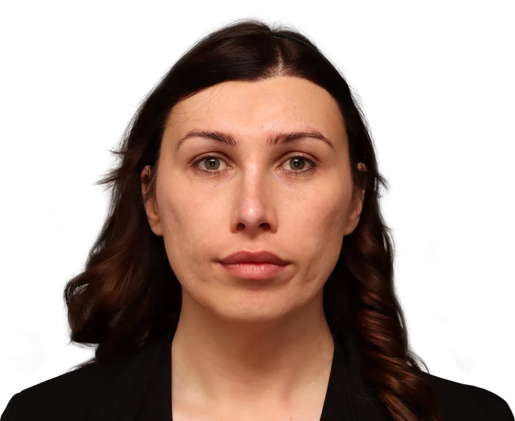 Close-up portrait of a woman with brown wavy hair and a neutral expression, wearing black, against a black background.