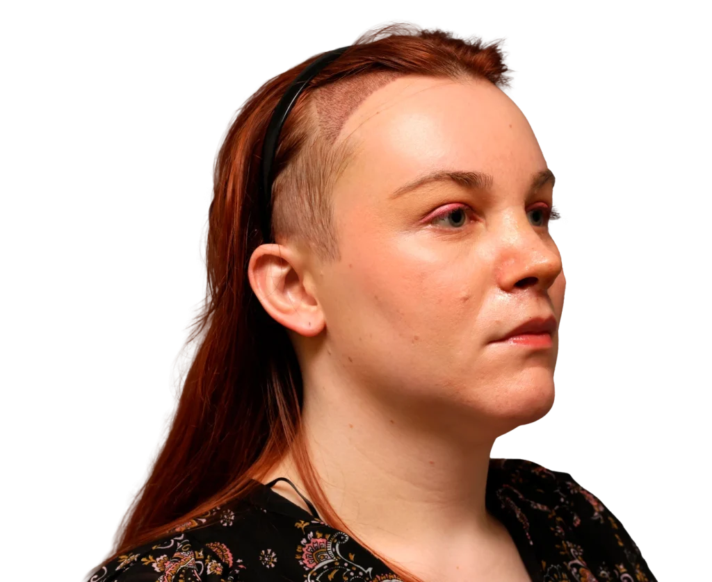 Side profile of a woman with a shaved temple, red hair, and a black floral blouse with a headband.