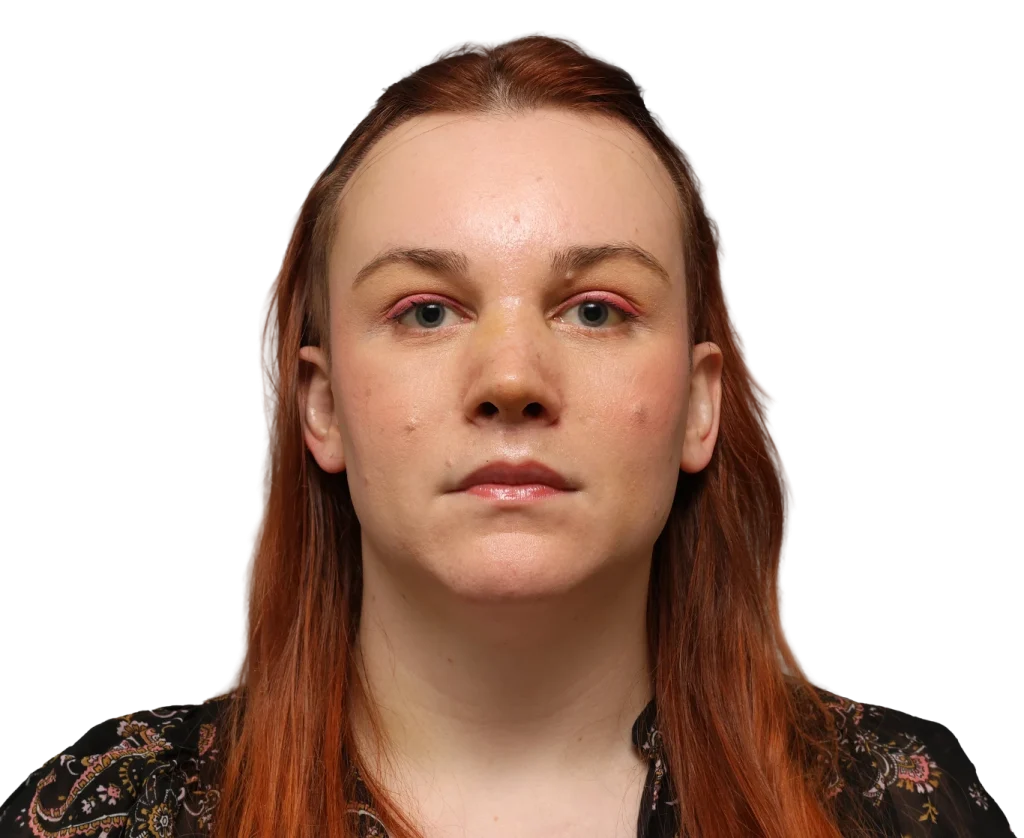 Front-facing headshot of a woman with red hair and pink eyeshadow, looking straight ahead against a black background.