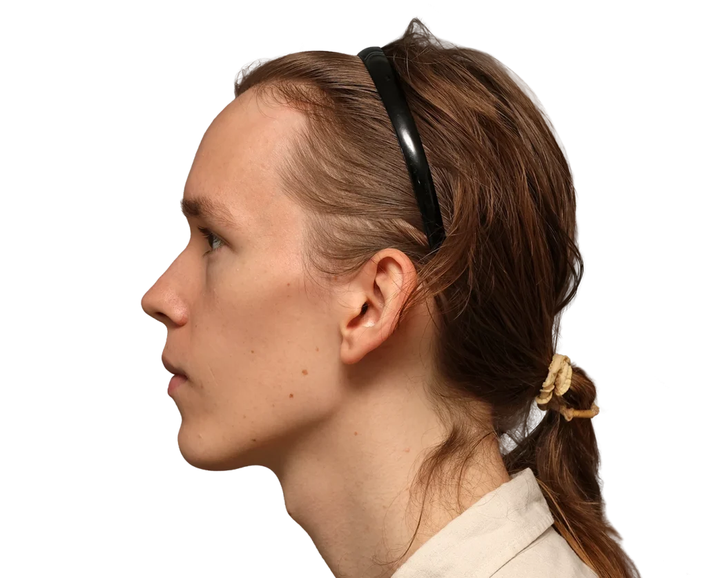 Side profile of a young woman with light brown hair tied back in a beige scrunchie, wearing a black headband against a dark background.